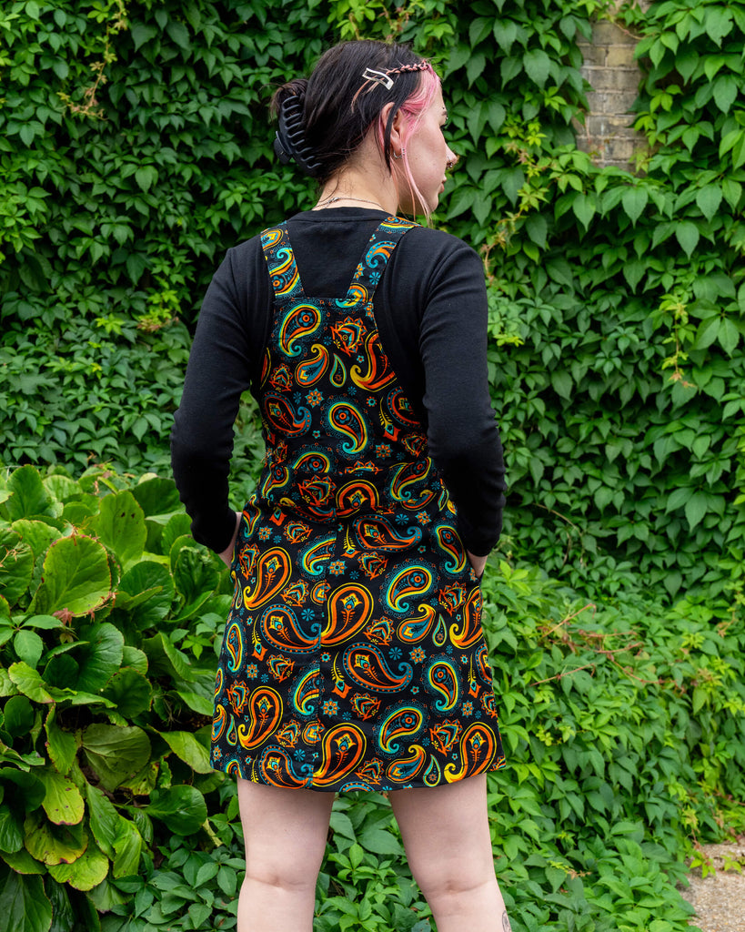 Rear view of a model in a Run & Fly slow fashion black paisley pinafore dress, complemented by a black top and chunky boots, standing in front of a green leafy wall.