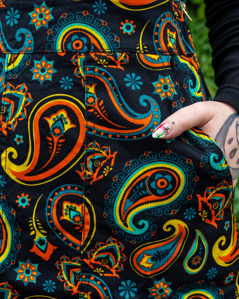 Close-up of Run & Fly's ethically made black paisley pinafore dress, highlighting colourful orange, teal, and yellow paisley patterns, with the model's hand casually in the pocket.