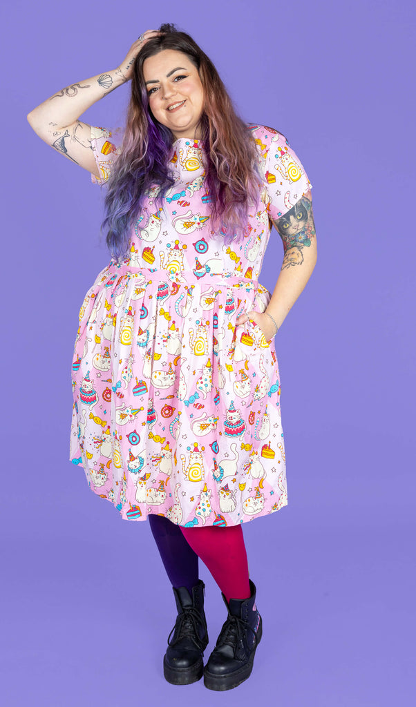Luisa, a femme model with purple hair and tattoos, is wearing Run & Fly x The Mushroom Babes Party Cats Stretch Belted Tea Dress with Pockets with pink and purple tights and black boots. The baby pink dress has an all over print of cats with party hats and clown makeup on, and various sweets. Luisa is stood in front of a purple backdrop and is smiling toward the camera with one hand in her hair and the other in the dress pocket.  