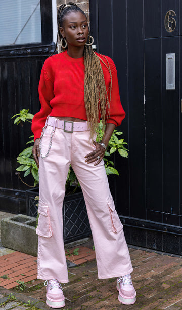 Yolande is stood outside in front of a black door wearing the 90's Baby Pink Wide Leg Oversized Stretch Cargo Trousers with a pink chain belt, red crop jumper and pink platform boots. She is facing forward leaning onto one hip with one hand in the side pocket, the other hand by her side, whilst looking off to the right. The trousers are an oversized fit, 2 flap cargo pockets on each leg as well as side pockets and back patch pockets.