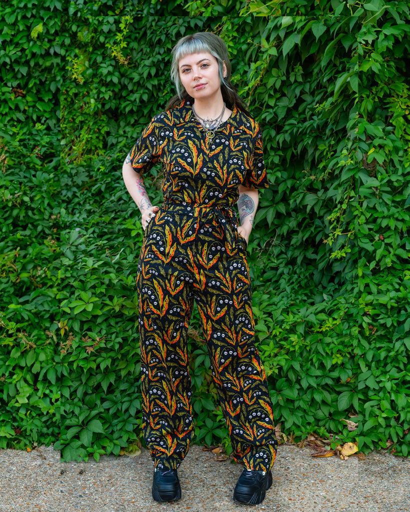 A person in a Run & Fly jumpsuit with skull and leaf prints, standing against a lush green backdrop. The playful print features vibrant oranges and greens, promoting ethical and slow fashion by Run & Fly.