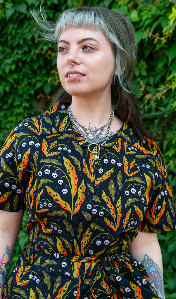 A person models a Run & Fly jumpsuit, featuring a bold pattern of white skulls and orange-green leaves on a black background. The person has light grey-green hair, a septum piercing, and silver layered necklaces, posing against a backdrop of greenery. Ethical slow fashion by Run & Fly, highlighting alternative, vibrant style.