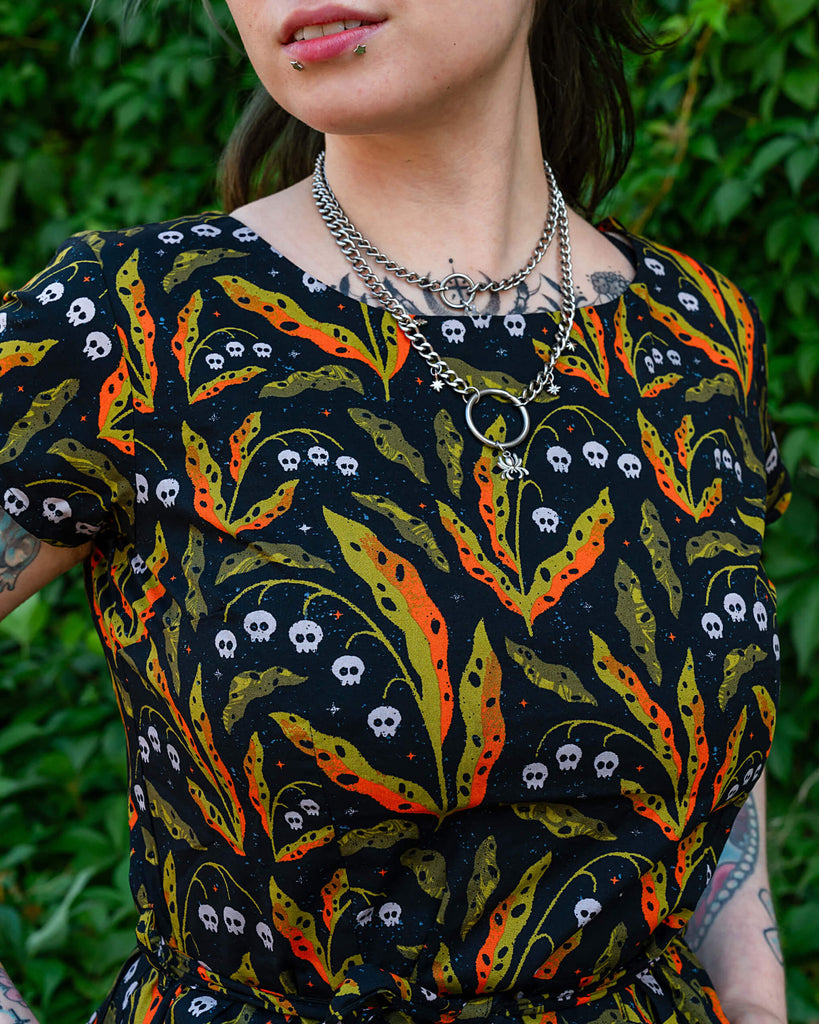 A close-up portrait of a person wearing a Run & Fly dress featuring a black base with orange, green, and white skull patterns. The person’s silver necklaces and piercings complement the outfit, with their hair blowing slightly in the wind. Run & Fly is an ethical slow fashion brand.