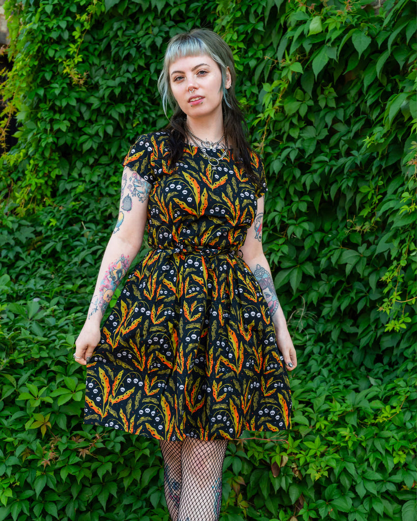 A person wearing a Run & Fly dress with a black base and a bright pattern of orange and green leaves, accented with small white skulls. They are posing in front of greenery, styled with fishnet tights and black platform shoes. Ethical slow fashion brand, Run & Fly.