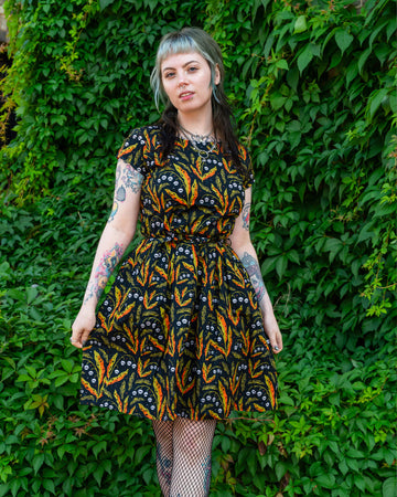 A person wearing a Run & Fly dress with a black base and a bright pattern of orange and green leaves, accented with small white skulls. They are posing in front of greenery, styled with fishnet tights and black platform shoes. Ethical slow fashion brand, Run & Fly.