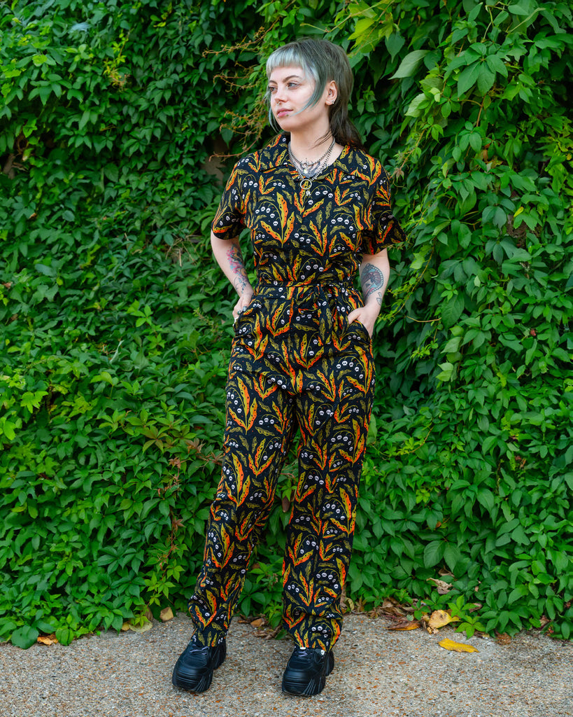 A person in a Run & Fly jumpsuit with skull and leaf prints, standing against a lush green backdrop. The playful print features vibrant oranges and greens, promoting ethical and slow fashion by Run & Fly.