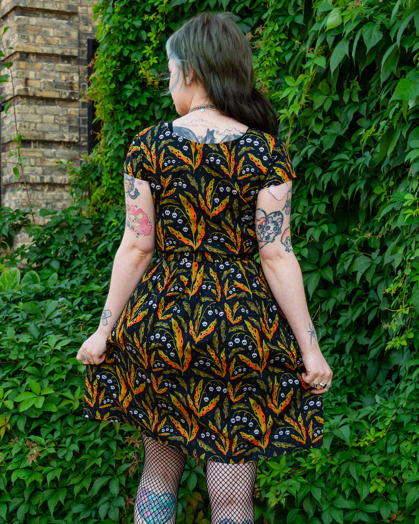 A person models the back of a Run & Fly dress featuring an orange and green leaf pattern with small skulls on a black background. The model’s tattoos and fishnet tights are visible, adding an alternative edge to the look. Ethical slow fashion by Run & Fly, promoting bold and quirky designs.
