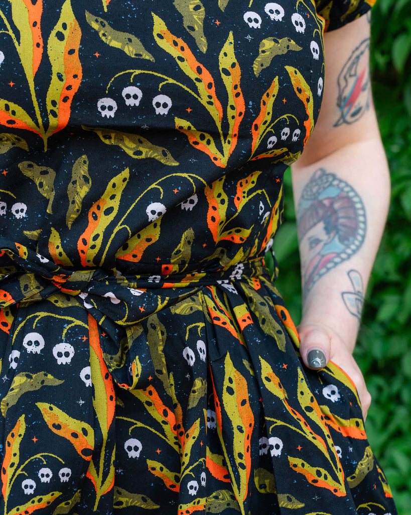 A close-up image of a Run & Fly dress showing the intricate skull and leaf print in bright orange and green hues on a black background. A silver necklace adds to the bold aesthetic of the ethical fashion brand.