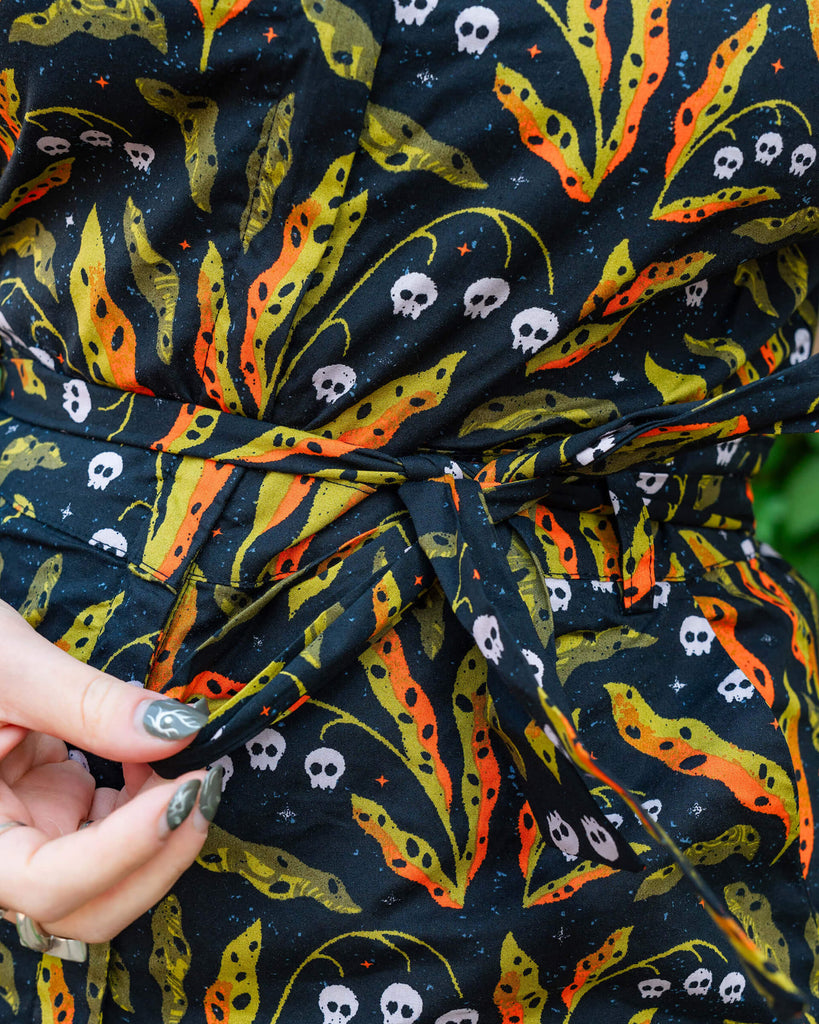 A detailed view of a vibrant Run & Fly jumpsuit featuring a black background with skulls, neon green and orange plant patterns. The fabric belt is shown being held at the waist by a person with black and white nail art. Ethical slow fashion brand, Run & Fly.