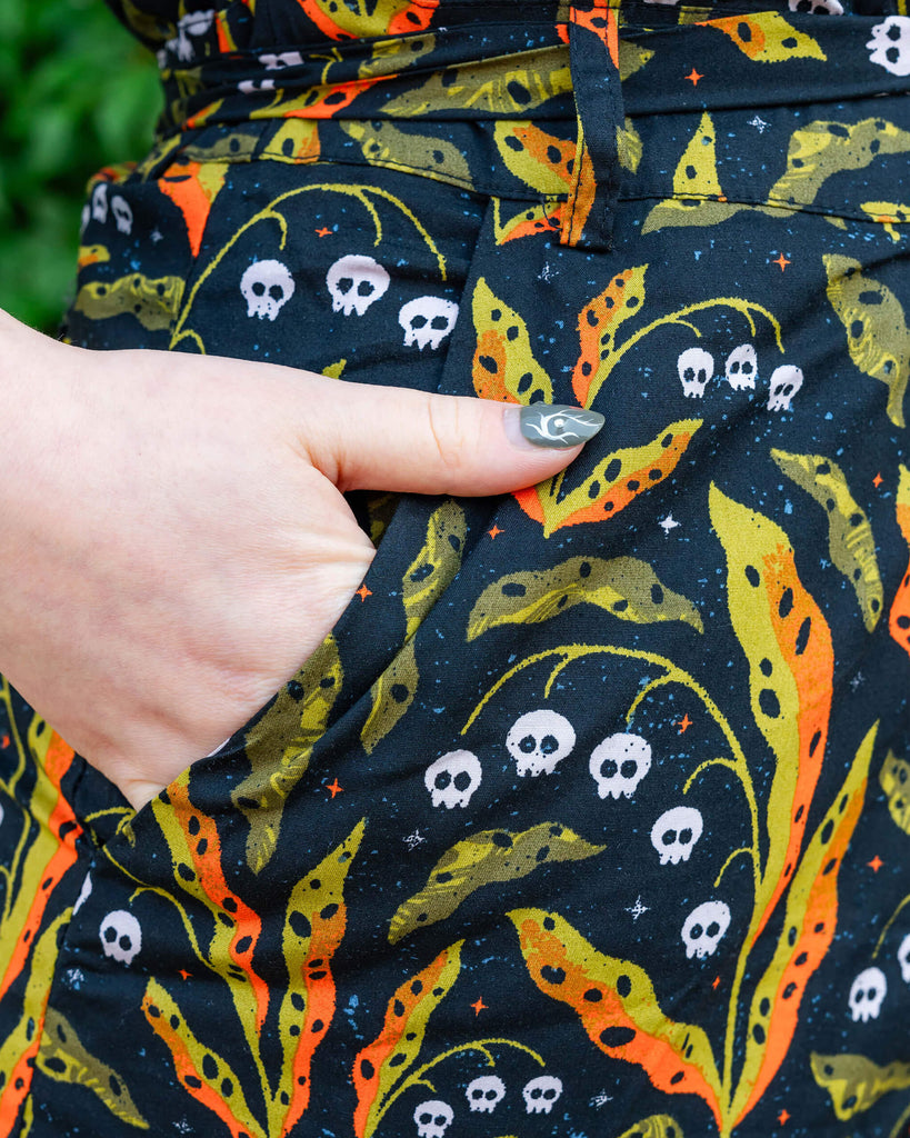 A person places their hand in the pocket of a Run & Fly jumpsuit. The fabric features a bold black background with bright orange and green leaf patterns interspersed with white skulls. The detailed shot highlights the practical pocket design, promoting Run & Fly's ethical and slow fashion focus.