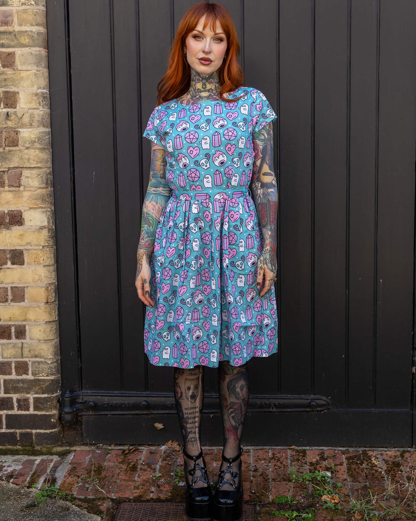 Front-facing image of a red-haired woman in a quirky blue dress featuring a pattern of ghosts, hearts, and pentagrams. She stands confidently in front of a dark door, showcasing her tattooed arms. The dress is from Run & Fly, a brand committed to ethical and sustainable fashion.