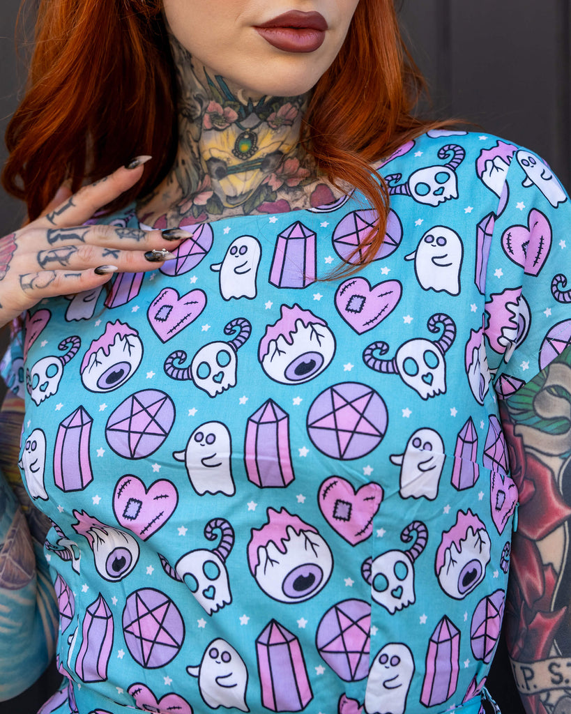 Red-haired woman with bold tattoos stands with her back to the camera, wearing a Run & Fly blue dress with a fun and spooky pattern featuring ghosts, pentagrams, and crystals. The brand is known for its ethical approach to slow fashion.