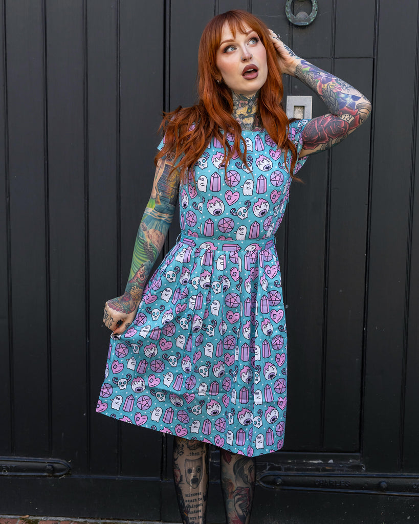 Full-length photo of a red-haired woman posing in front of a black wooden door, showcasing her colourful tattoos and wearing a playful blue dress with a ghost and crystal print by Run & Fly. The brand is celebrated for its ethical and slow fashion practices.