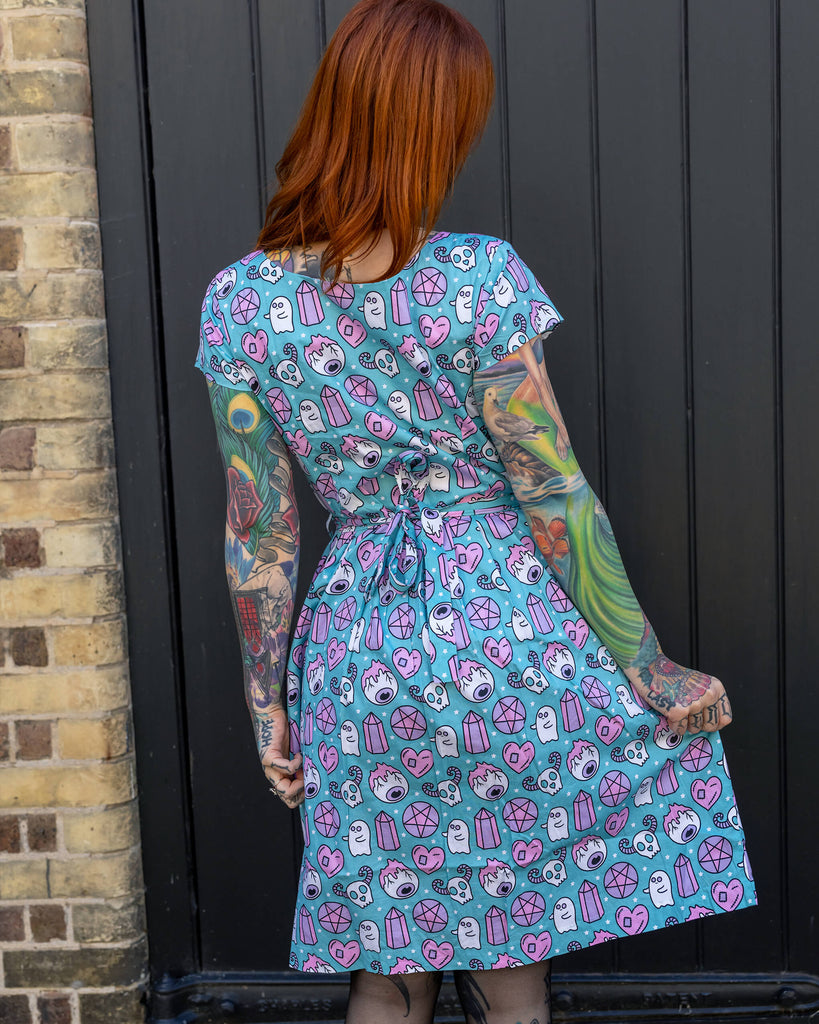 Red-haired woman with bold tattoos stands with her back to the camera, wearing a Run & Fly blue dress with a fun and spooky pattern featuring ghosts, pentagrams, and crystals. The brand is known for its ethical approach to slow fashion.