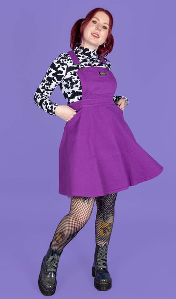 model with red hair wearing a purple flared pinafore paired with a long sleeve cow print turtle neck, fishnet tights and boots. Model is smiling with her hands in the pinafore pockets in front of a purple background.