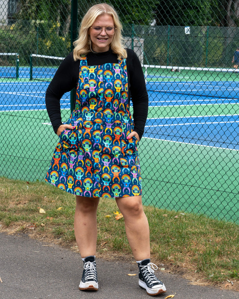 Full-body shot of Betty modelling the Run & Fly Rainbow Frogs Stretch Twill Pinafore Dress. The A-line mini silhouette features an all-over rainbow frog print with adjustable buckle straps and front pockets. Styled with a black top and black Converse trainers, highlighting the playful and ethical slow fashion piece.