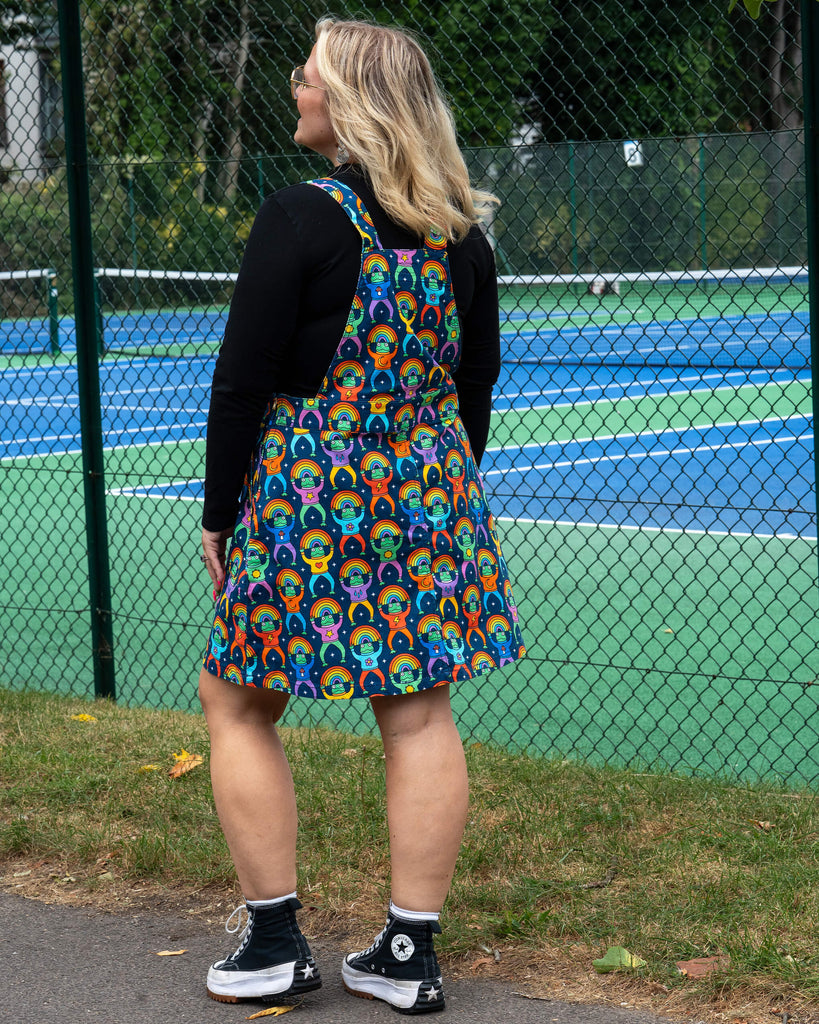 Back view of Betty wearing the Run & Fly Rainbow Frogs Stretch Twill Pinafore Dress. The pinafore shows off the rainbow frog all-over print on stretch twill fabric, styled with a black top and Converse trainers. A quirky and colourful ethical slow fashion outfit.