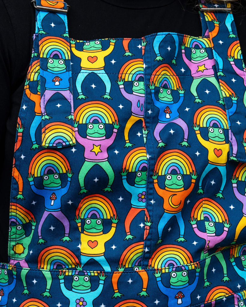 Close-up of the Run & Fly Rainbow Frogs Stretch Twill Pinafore Dress fabric. The design by Wafflemama features colourful cartoon frogs holding rainbows, wearing jumpers with stars, hearts, mushrooms and moons. Ethically made stretch twill fabric with bold, fun detailing.