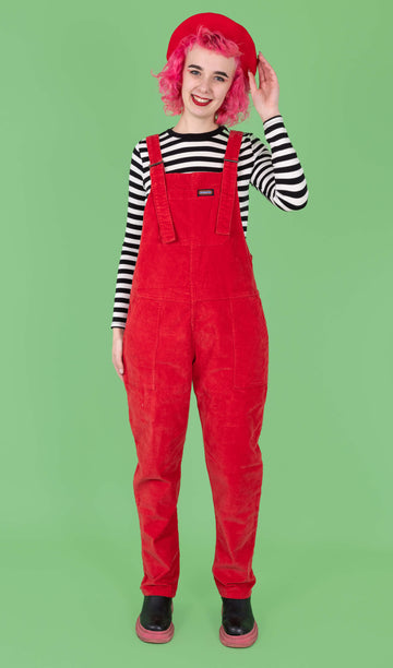 A pink haired femme model wearing the run and fly red stretch corduroy dungarees with a red beret, black boots and a black and white stripe long sleeve tshirt. They are facing forward to highlight the retro roller straps and front bib pocket with a rainbow run & fly label. They are smiling to camera with one hand resting by their side whilst the other rests on the beret.