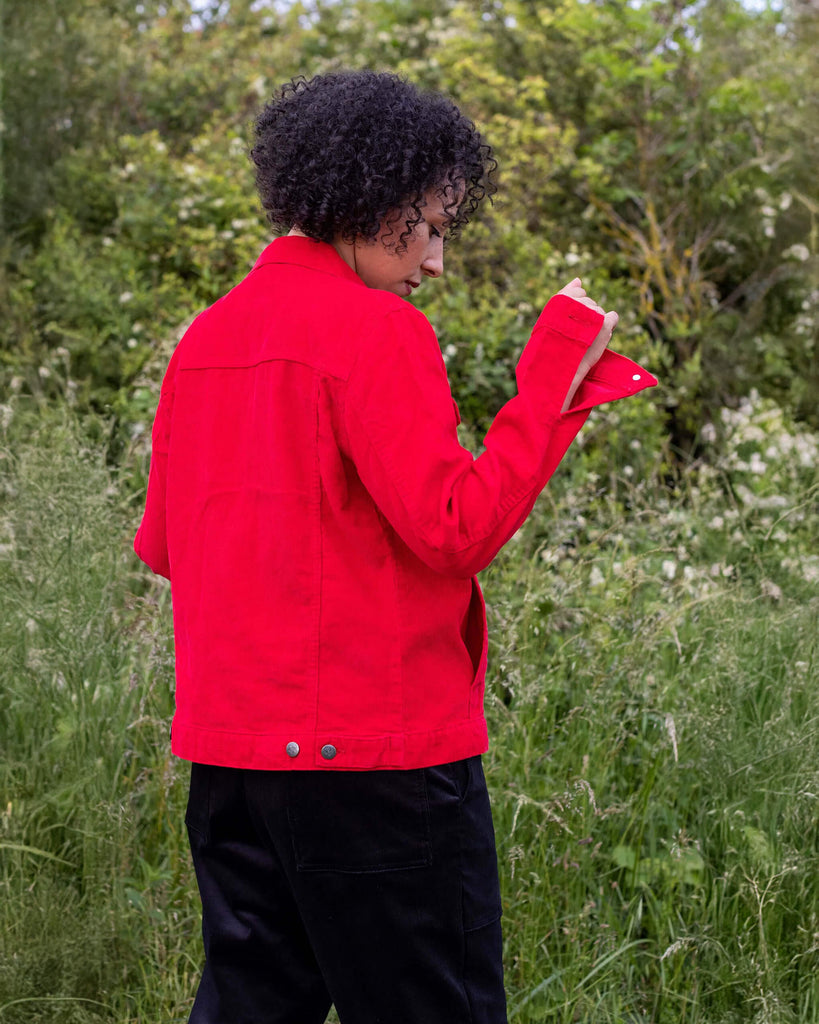 Amy is stood outside in a field area wearing the retro vintage red cord unisex western jacket with black dungarees. They are facing away from the camera with one arm raised up to show off the cuff detailing. Photo is cropped from the hips up.
