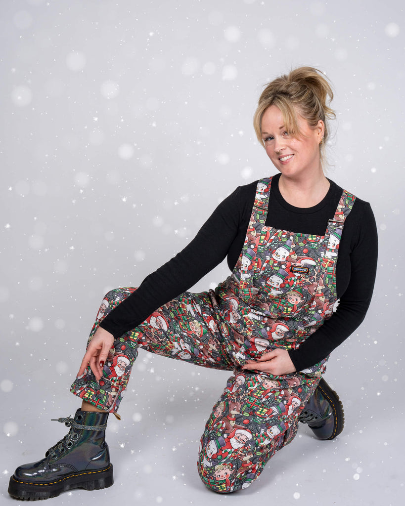 Karina kneels on one knee, wearing Run & Fly Christmas dungarees adorned with colourful retro holiday characters. The snowy overlay adds a magical touch, enhancing the festive atmosphere of the shoot.