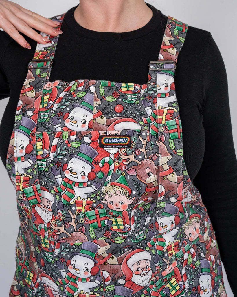 A close-up shot of a festive Run & Fly dungarees top featuring an all-over print of retro Christmas characters, including Santa, reindeer, elves, and snowmen, paired with a black long-sleeve top. Model Karina smiles subtly, showcasing the detailed and vibrant holiday-themed design.