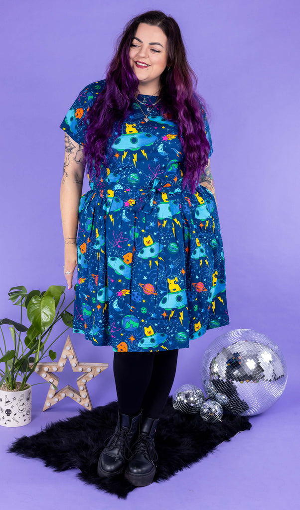 Luisa, a femme model with purple hair and tattoos, is stood in a photography studio in Hove in front of a purple backdrop wearing Dogs in Space Stretch Belted Tea Dress with Pockets with black tights and boots. The print features orange and yellow bull terriers, pomeranians and labradors in blue spaceships on a space themed blue background with stars, planets and rockets. The dress is short sleeved with pockets and a belt tie. Luisa is facing the camera and looking off to one side smiling.