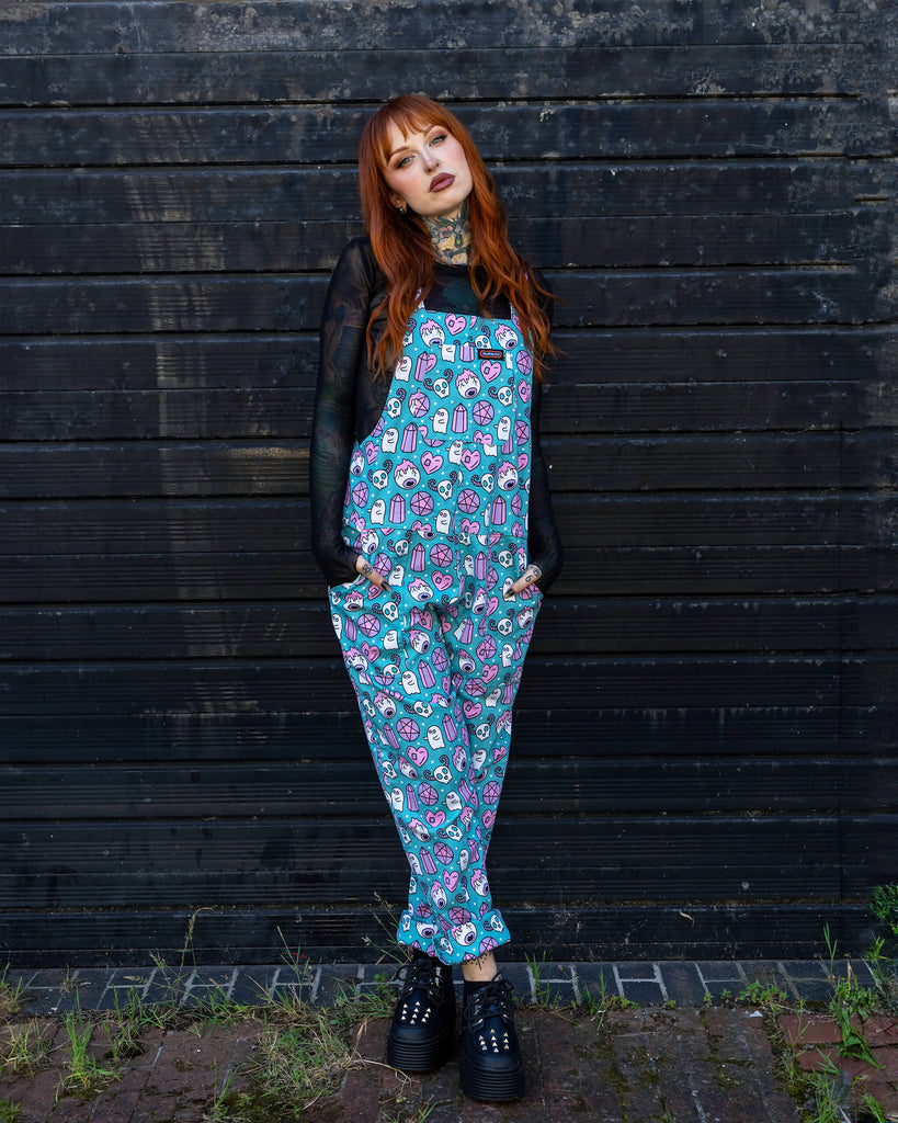 The woman poses confidently in the full outfit of turquoise Run & Fly dungarees, decorated with a spooky-themed print in pastel shades. The outfit is paired with a black mesh top and chunky black platform shoes, set against a dark wooden background, creating a striking contrast.