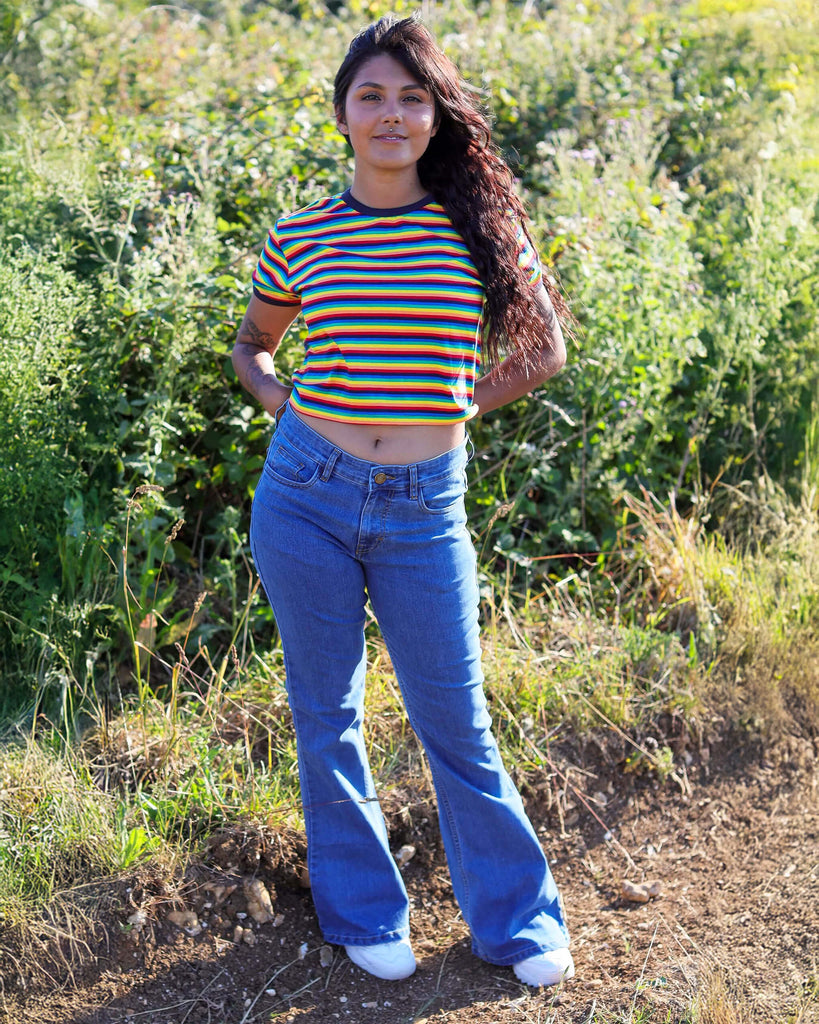 Meera is stood in a grass field wearing the stone wash blue stretch denim rock n roll bell bottom flares with a retro rainbow stripe short sleeved tshirt and white trainers. She is facing forward posing leaning on one leg with both hands resting behind her and smiling to camera.