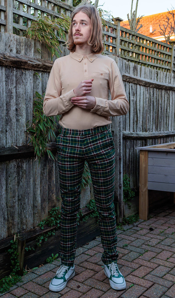 Jack is stood in a garden wearing the retro mod stretch plaid green tartan slim skinny fitting trousers with a sand coloured long sleeve shirt with white trainers. They are facing the camera posing doing up one of the shirt cuff buttons and looking off to the left.