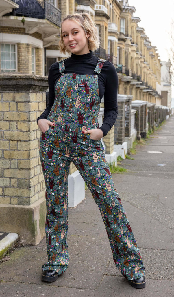 Model with short blonde hair wearing Peace and Love Stretch Skinny Flared Dungarees paired with long sleeve black turtleneck top and chunky black boots. The dungarees are an olive green colour with hands in different skin tones making the peace sign, various flowers, peace symbols and love hearts in white, yellow, purple and pink and 'peace' and 'love' printed on. There is a Run&Fly logo on front pocket on chest of dungarees. Model is posing with hands in pockets and smiling at camera.