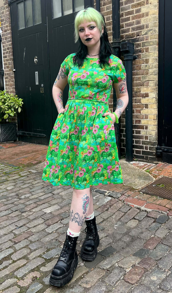 Tattooed model with green and black hair and dark makeup wearing Run & Fly x The Mushroom Babes Frogs Stretch Belted Tea Dress with Pockets, paired with black chunky boots. The dress is a bright green colour with an all over pattern of green frogs and lily pads, blue puddles, pink and orange flowers and mushrooms and various insects. Model is posing with one foot in front of other with hands in dress pockets.