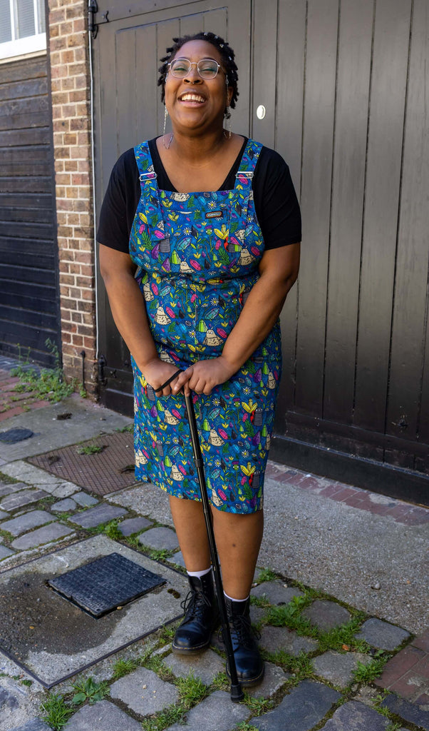 Han, a black femme model with silver glasses with a chain, is stood outside in Hove in a mews wearing Run & Fly x Katie Abey Crystal Critters Stretch Twill Long Pinafore Dress with a black t shirt underneath and black boots. The blue pinafore dress has an all over print of critters and crystals illustrated by Katie Abey. Han is smiling toward the camera and is leaning on her black walking aid.
