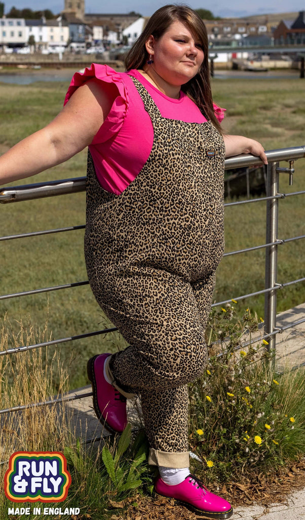 Nat is stood near a river wearing the natural leopard print stretch twill dungarees with a bright pink ruffle short sleeve tshirt underneath and bright pink brogues. They are posing leaning back on the metal railing with both hands resting up beside them and one foot resting back on the railing.