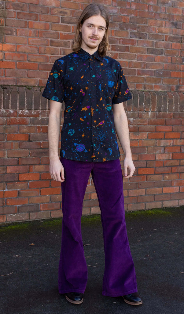 Jack is stood in front of a brick wall wearing the purple corduroy bell bottom flares with the cosmic space shirt and black boots. They are facing forward posing with both arms resting by their side and smiling to camera.