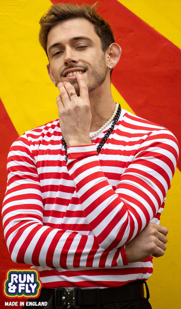Zach is stood in front of a red and yellow painted wall wearing the red and white stripe long sleeve tshirt with black trousers. He is facing forward posing with one arm across his chest whilst the other hand is resting across on his chin. Photo is cropped from the hips up.