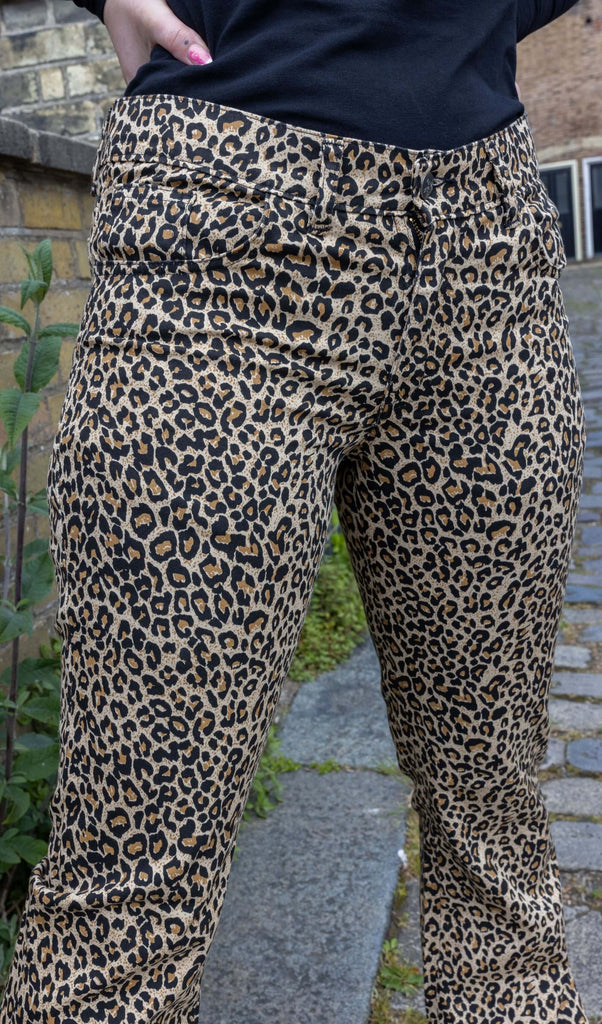 Close up of model wearing Leopard Print Bell Bottom Stretch Super Flares, paired with a long sleeve black turtleneck top. The flares are a natural leopard print pattern and colour. The close up shows the silver button on the flares as well as belt loops and two front pockets. Model is posing with hand on hip and pink nail polish.