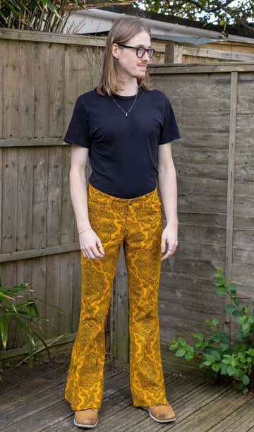 Jack, a white male model with long hair and glasses is wearing Gold Hendrix Paisley Corduroy Retro Bell Bottom Flares paired with a black t-shirt and brown shoes. The trousers are a yellow gold colour with an all over black paisley pattern. Jack is stood outside in East Sussex in front of a wooden fence facing the camera.