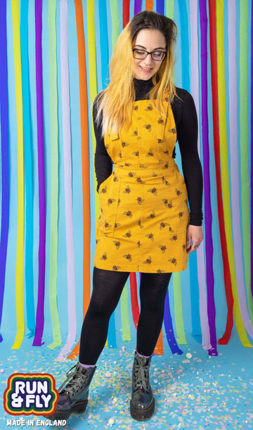 Jojo is stood in front of a blue rainbow studio background wearing the bees knees bee print gold stretch twill pinafore dress with a long sleeve black tshirt and grey boots. She is facing forward leaning back on one leg with one hand in the pinafore pocket whilst smiling and looking down.