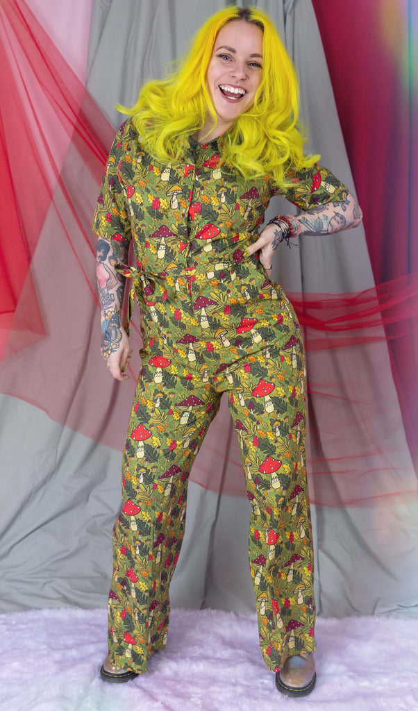 Katie Abey is stood in a photography studio in Hove wearing Run & Fly x Katie Abey Mushroom Boi's Jumpsuit. The jumpsuit has an all over 'mushroom bois' print illustrated by Katie Abey. Katie is smiling and facing the camera with her hand on her hip.