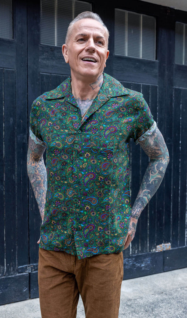 white male heavily tattooed model wearing Forest Green Paisley Short Sleeve Rayon Shirt paired with brown corduroy trousers. The shirt is a forest green colour with an all over paisley print in various muted rainbow colours. The model is stood outside in Hove in front of a black door smiling. 