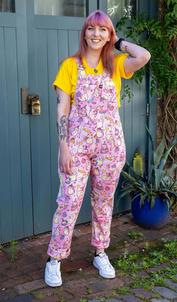Stacie is stood in a mews in Hove wearing Run & Fly x The Mushroom Babes Party Cats Stretch Twill Dungarees with a bright yellow top underneath and white trainers. The dungarees are baby pink with white cats with party hats on and eating birthday cake, as well as various pink, blue and yellow sweets designed by The Mushroom Babes. Stacie is facing the camera and smiling with one hand in her hair and other resting by her side.