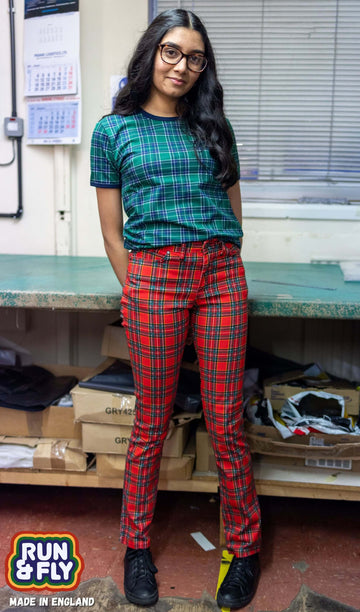 Model is stood in our warehouse area wearing the mid rise red tartan stretch skinny jeans with a green tartan tshirt and black trainers. They are facing towards the camera leaning against a desk with both hands behind their back and smiling.