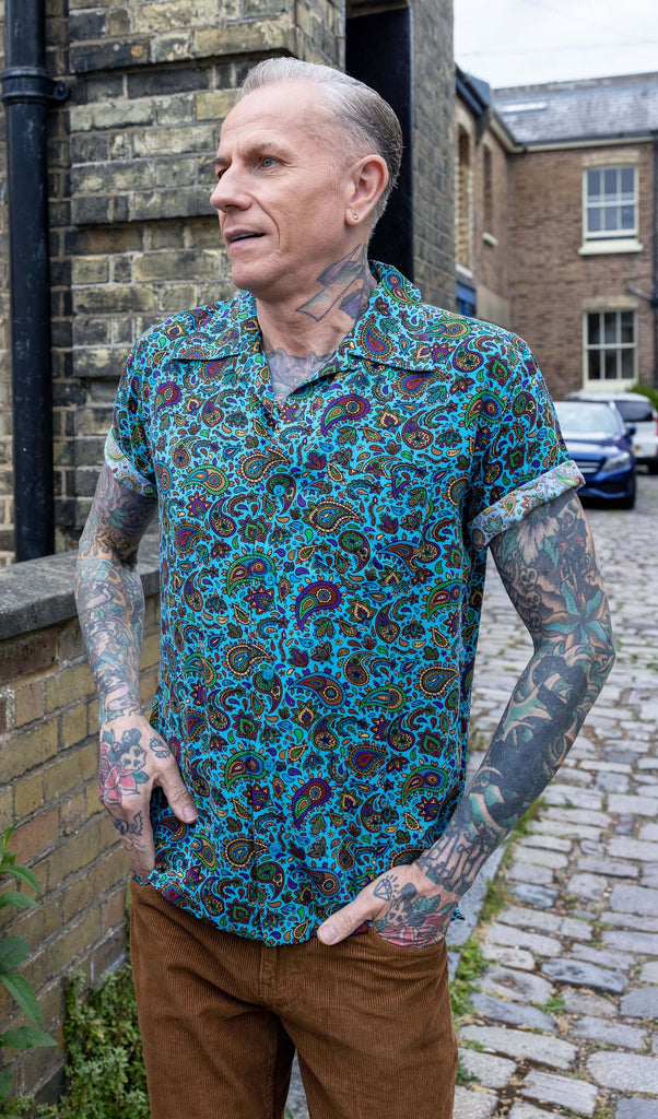 James is stood outside in a mews in Hove wearing wearing Sky Blue Paisley Short Sleeve Rayon Shirt paired with tan corduroy trousers. The collared, button up shirt is a sky blue colour with an all over paisley print in various muted rainbow colours. James is posing with his hands in his pockets and looking off to one side.