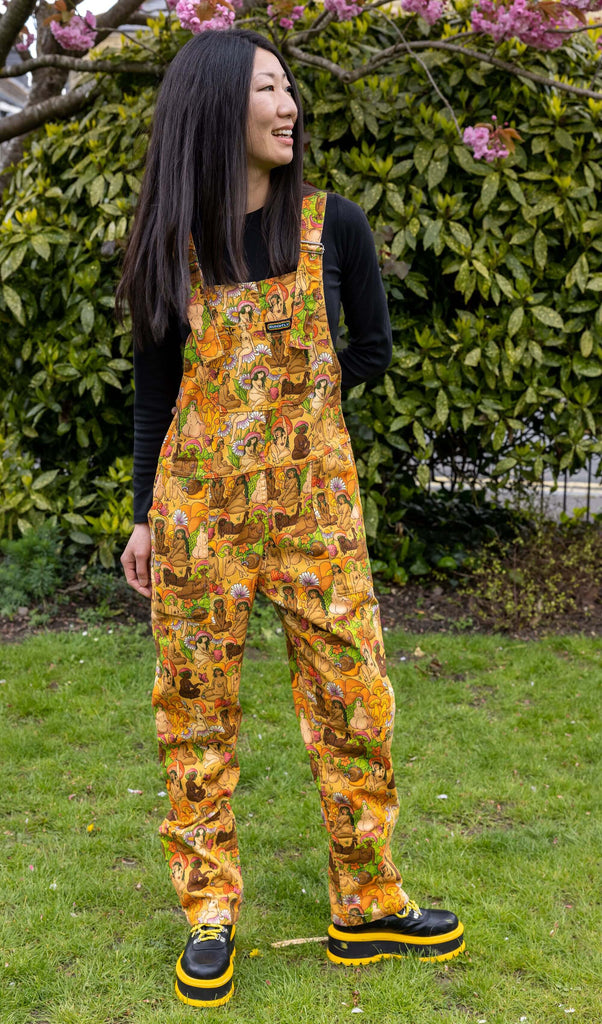 Arisa is standing in a garden, wearing the Run & Fly. The orange Mushroom Babes Natural Babes Stretch Twill Dungarees. Model is looking away from the camera and smiling with one hand by her side and one behind her back, dungarees worn with chunky platform boots and a black long sleeved top.