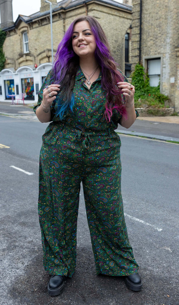 Model with blue, purple and pink hair is wearing Forest Green Paisley Jumpsuit paired with black boots. The jumpsuit is a forest green colour with an all over paisley print in various muted rainbow colours. There is a matching fabric belt tied around the model's waist. The model is stood outside in Hove smiling with her hands in her hair. 