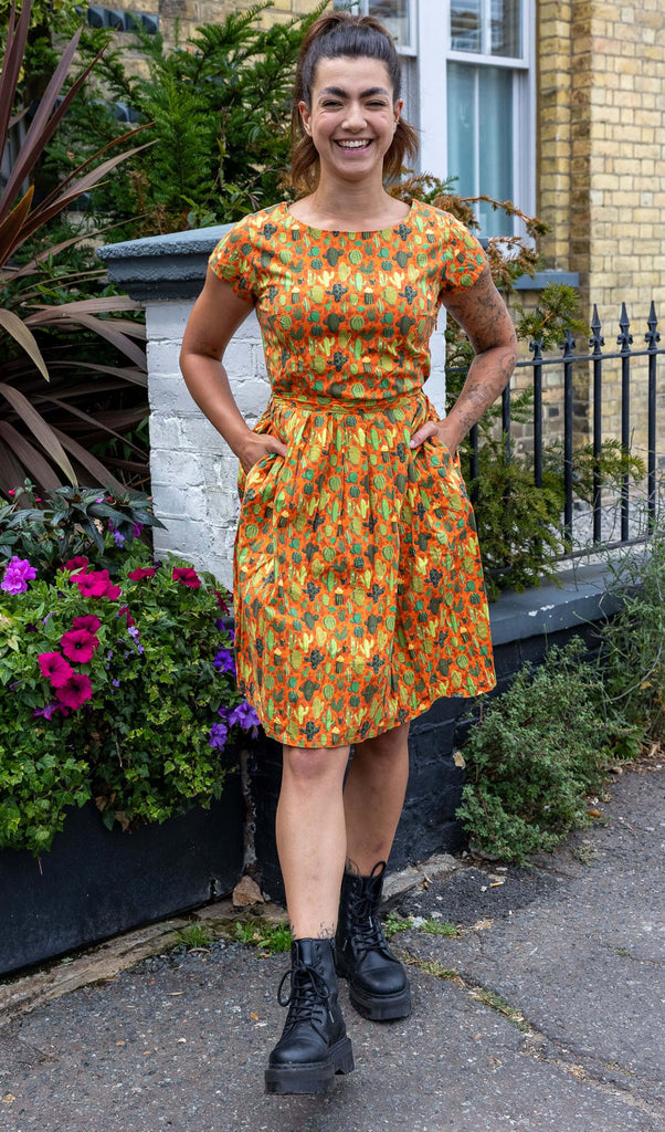 Model wearing Orange Cactus Tea Stretch Dress with Pockets paired with chunky black boots. The background colour of the dress is a bright orange with various cactus patterns on it in different shades of green. Model is smiling and has her hands in the dress pockets.