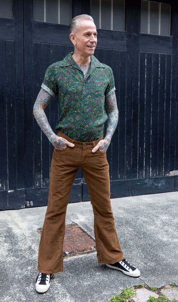 James, a tattooed male model, is stood outside in Hove in a mews wearing Tan Corduroy Bootcut Flares with Forest Green Paisley Short Sleeve Rayon Shirt. James is stood facing the camera with his hands in his pockets and looking off to one side smiling. 