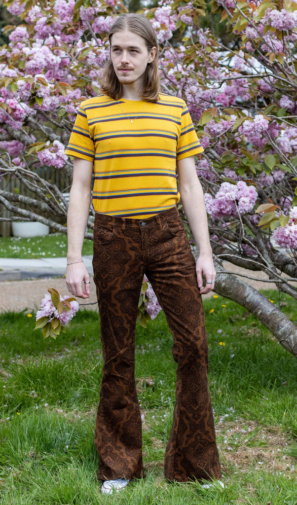 Jack, a male model with long blonde hair is stood outside in East Sussex by a blossom tree wearing Hendrix Tan Paisley Corduroy Retro Bell Bottom Flares paired with Gold Sunset Stripe Retro T Shirt. The trousers are a tan colour with an all over black paisley print. Jack is posing facing the camera with his arms down by his sides.