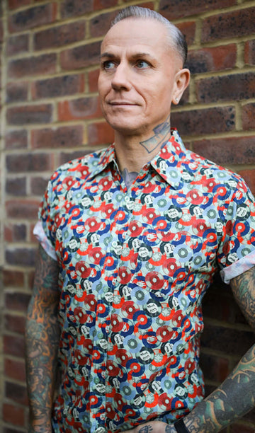 Jim is stood in front of a brick wall wearing the red and blue record short sleeve shirt with blue denim jeans. They are heavily tattooed with swept back silver hair. They are facing the camera posing with one hand on their hip whilst the other rests by their side. Photo is cropped from the hips up.